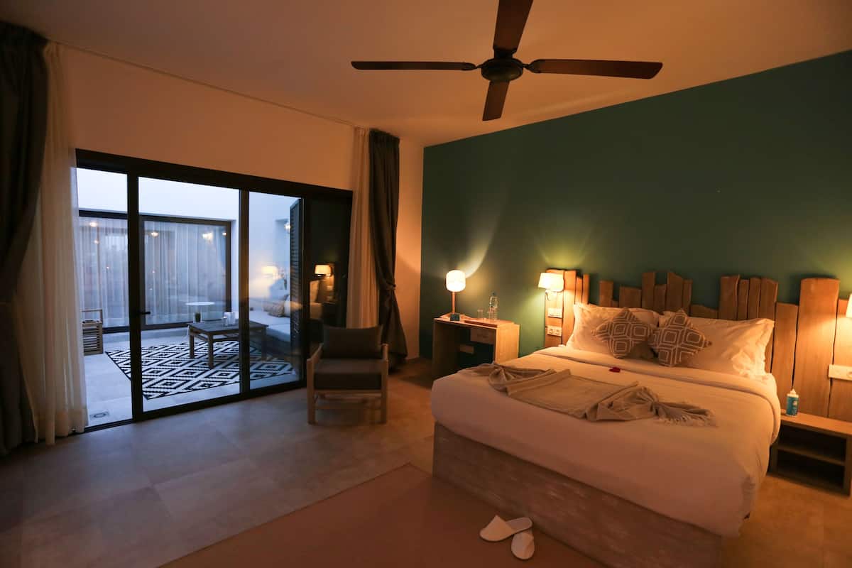 A hotel room with a bed, a ceiling fan, a nightstand, a chair, a table, and a view through a sliding glass door to an outdoor area.