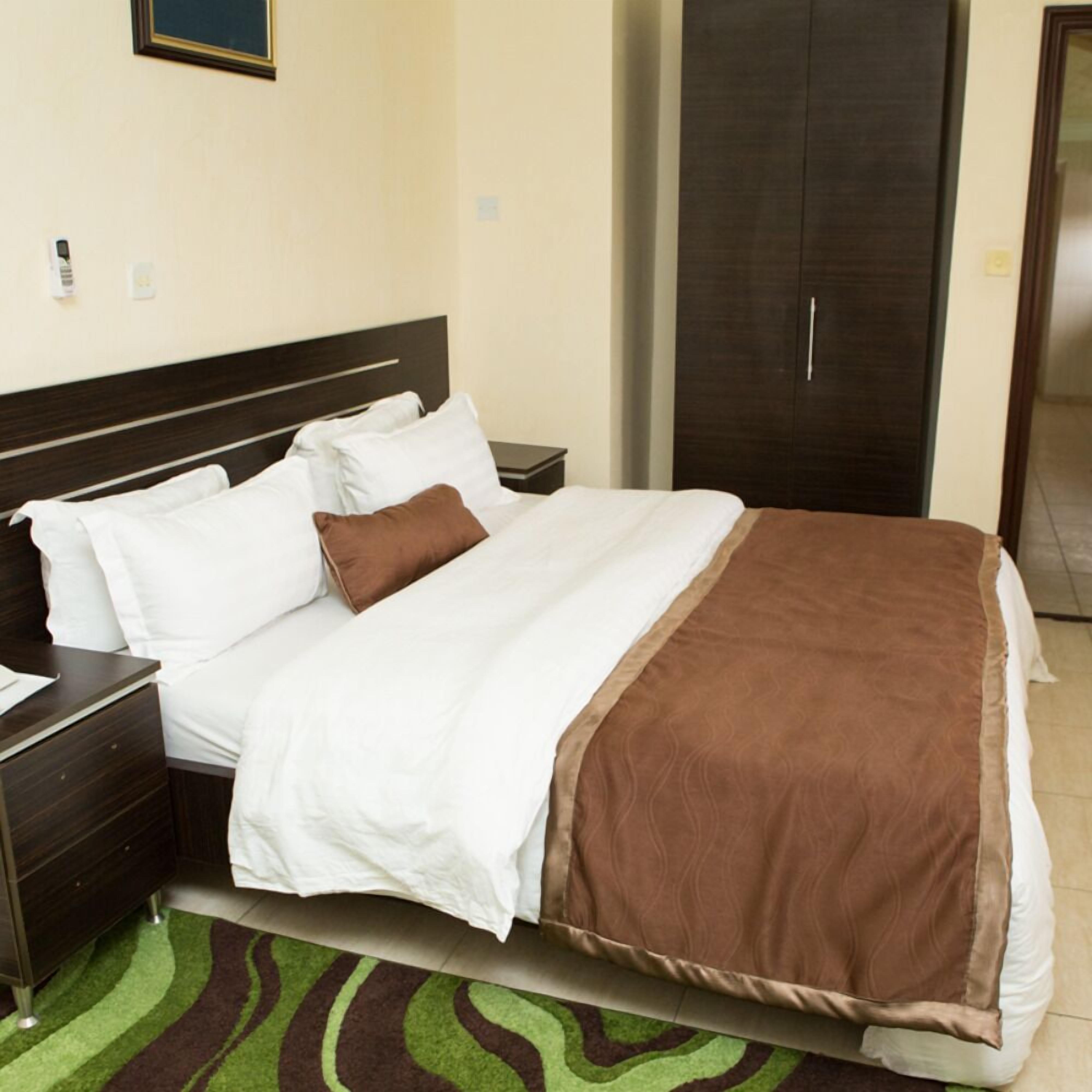 A neatly made bed with white and brown bedding, a dark wooden headboard, a nightstand with a lamp, and a green and brown striped rug.