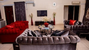 A living room with a red sofa, a grey sofa, and a brown sofa. There is a TV on the wall, a table with magazines, and two vases with flowers. - Ishakool Hotels (Abuja)