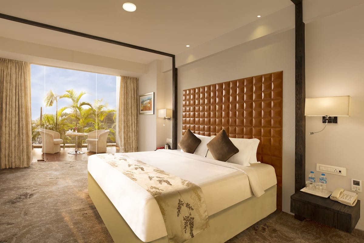 Une chambre d’hôtel avec un grand lit, un bureau avec une lampe, une chaise, une vue sur des palmiers et une fenêtre avec des rideaux.