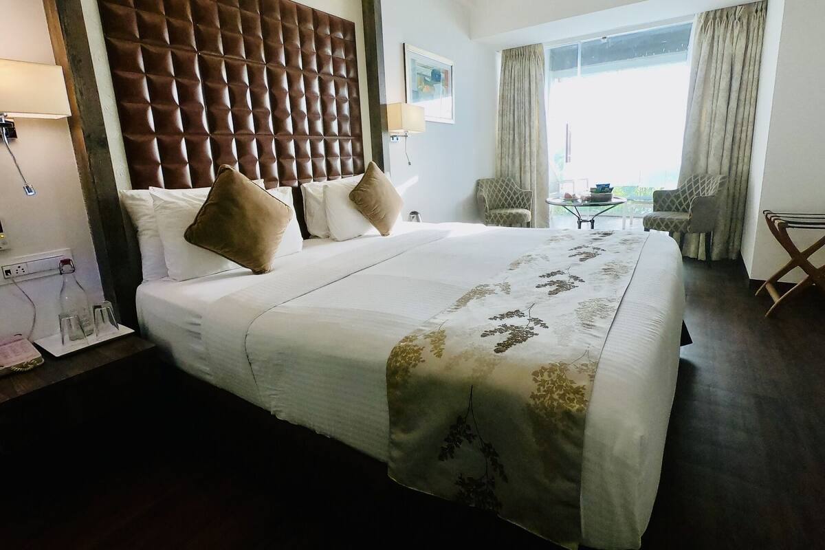 Une chambre d’hôtel avec un grand lit, une tête de lit en bois, deux chaises, une petite table et une fenêtre avec des rideaux.