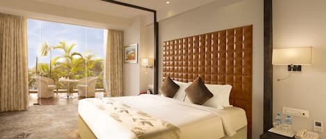 Habitación de hotel con una cama grande, una mesita con sillas, vista a palmeras y una ventana con cortinas.