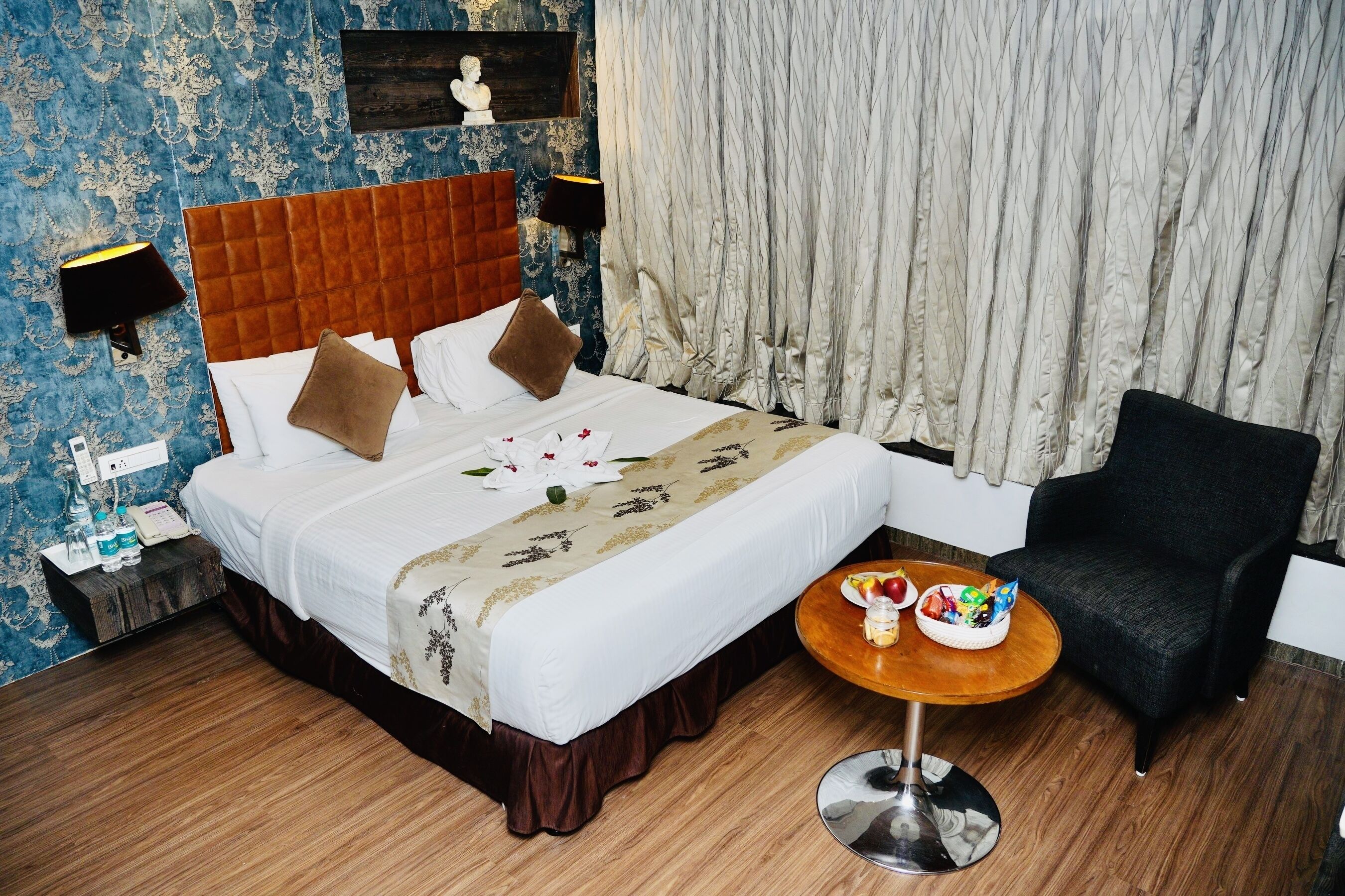 A hotel room with a large bed, a round wooden side table, a black armchair, and a wall with a patterned wallpaper.