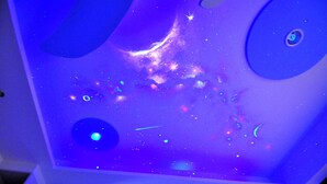 Una habitación con proyección de cielo estrellado en el techo, un ventanal circular y una pequeña lámpara circular.