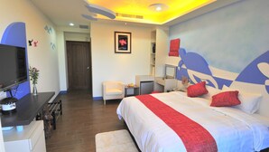 Una habitación de hotel moderna con una cama grande, un escritorio con televisor de pantalla plana, una silla y una pequeña mesa con un florero y fruta.