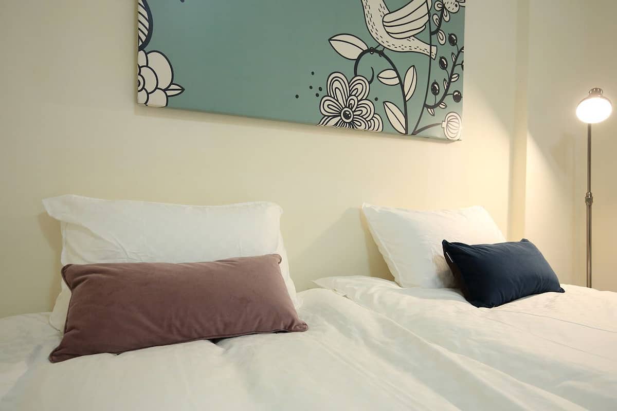 A hotel room with two beds, each with white bedding and different colored pillows. A floor lamp is on the right side of the room.