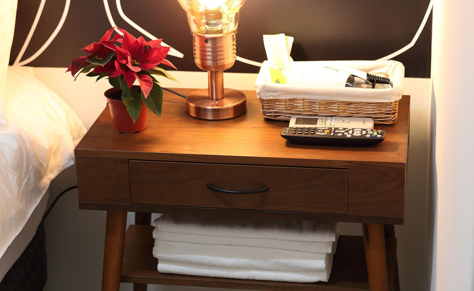 A wooden bedside table with a lamp, a remote control, a basket with tissues, and a potted plant.
