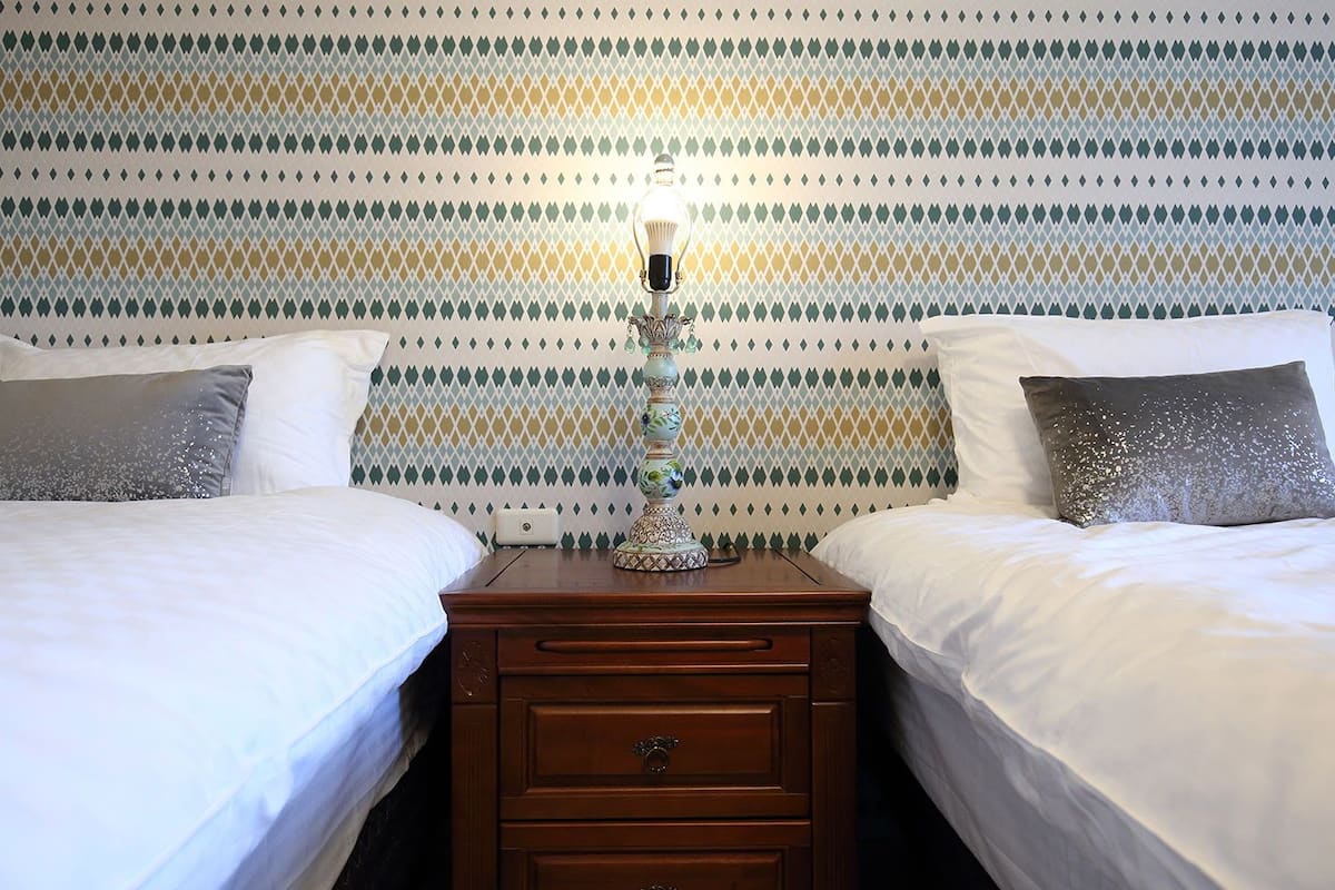 A hotel room with two beds, a nightstand with a lamp, and a patterned wallpaper.