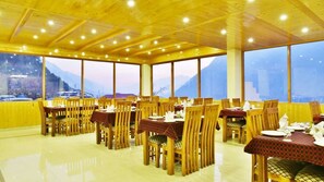 A restaurant with wooden tables and chairs, large windows, and a view of mountains. - Whistling Pines Resorts and Spa (Manali)