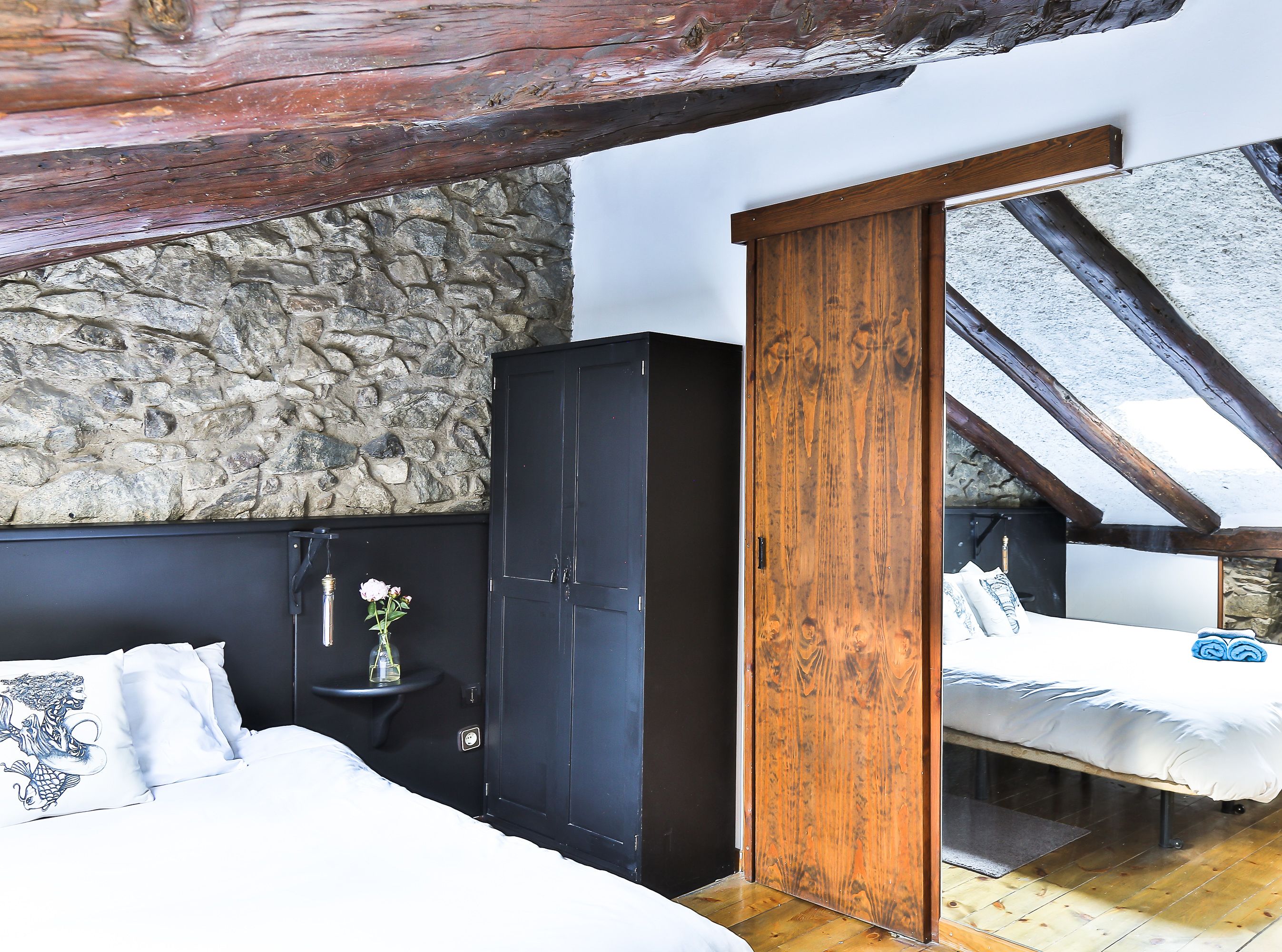 Un dormitorio con pared de piedra, vigas de madera, cama, armario y una pequeña mesa con un jarrón.
