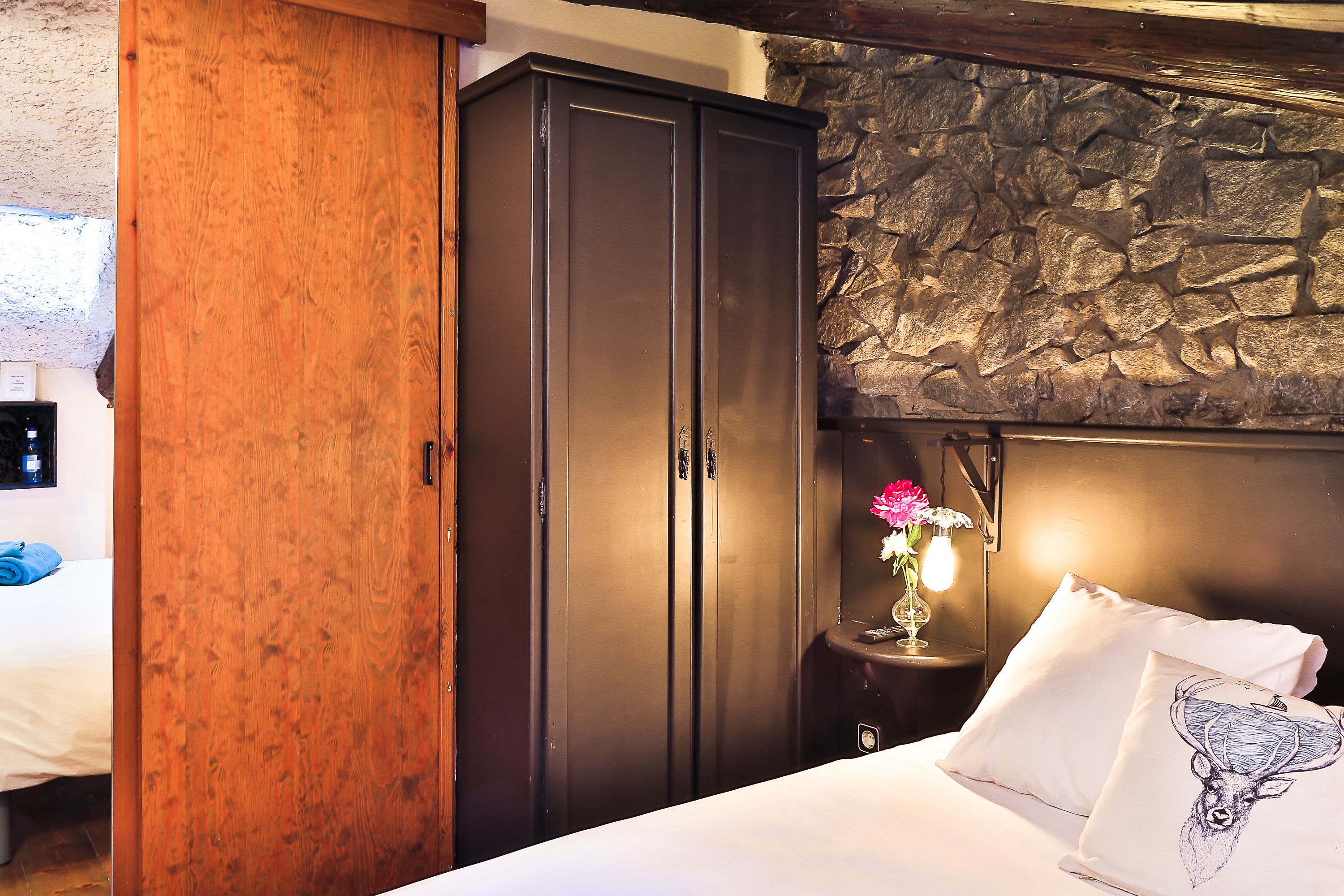 A bedroom with a wooden wardrobe, a stone wall, a bedside table with a lamp, and a bed with a pillow featuring a deer design.