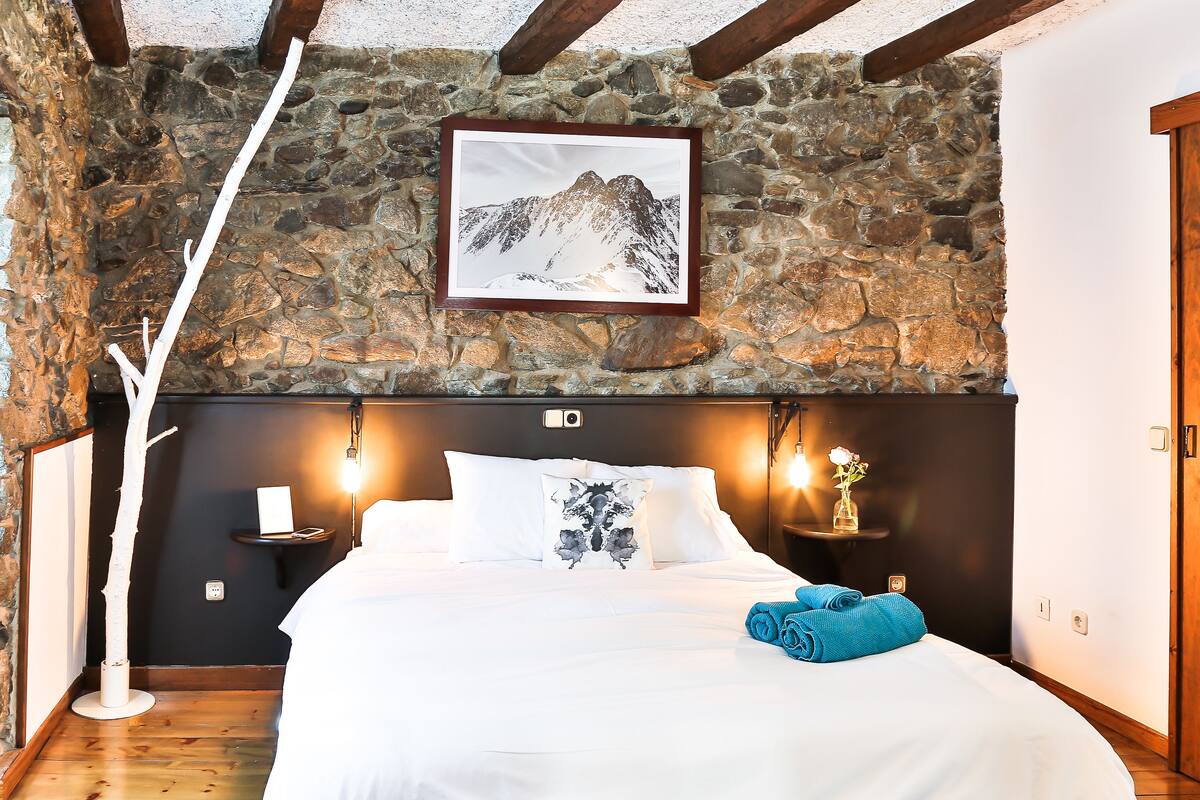 A bedroom with a stone wall, a framed picture of mountains, a bed with white bedding, and a blue towel.