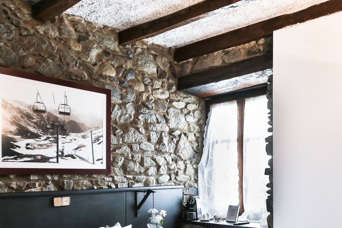 A bedroom with a stone wall, a bed with white bedding, a framed picture, and a window with curtains.
