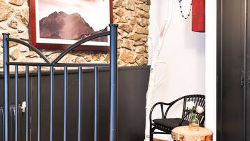 Una habitación con pared de piedra, cama con toalla enrollada, una silla, una mesita con un florero y una puerta.