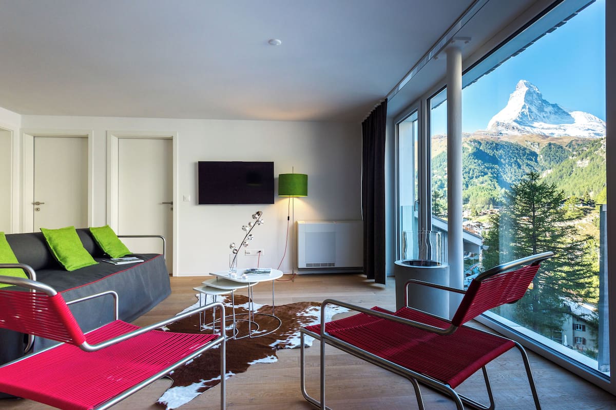 A modern hotel room with a large window offering a mountain view, a sofa with green cushions, a red chair, and a small table with a lamp.