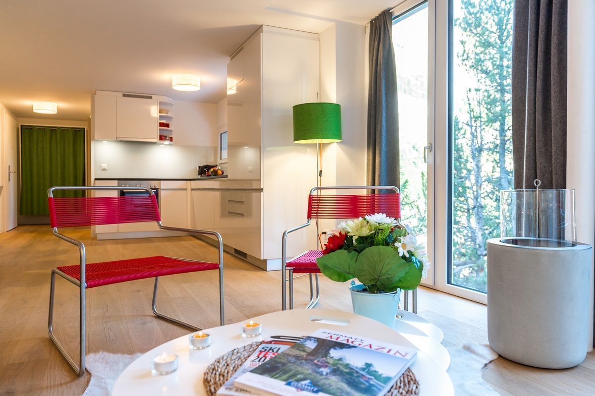 A modern room with a dining table, two red chairs, a green lamp, a vase with flowers, and a large window with curtains.