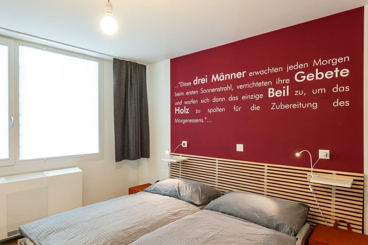 A bedroom with a large window, a bed with grey bedding, a red accent wall with a quote, and a bedside table with a lamp.
