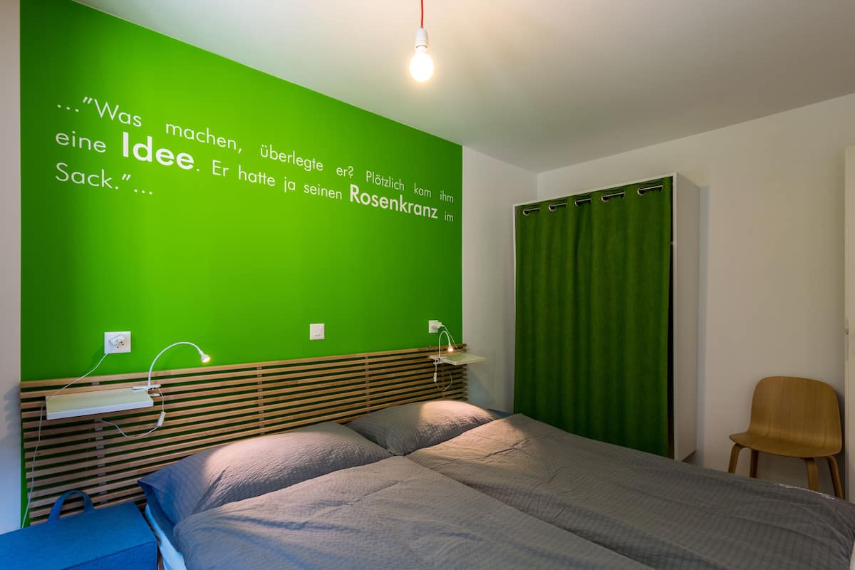 A bedroom with a green accent wall, a wooden headboard, a bed with grey bedding, a bedside table with a lamp, and a green curtain.