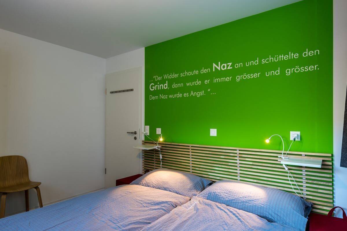 A bedroom with a green wall, a wooden headboard, a bed with blue bedding, a chair, and a bedside table with a lamp.