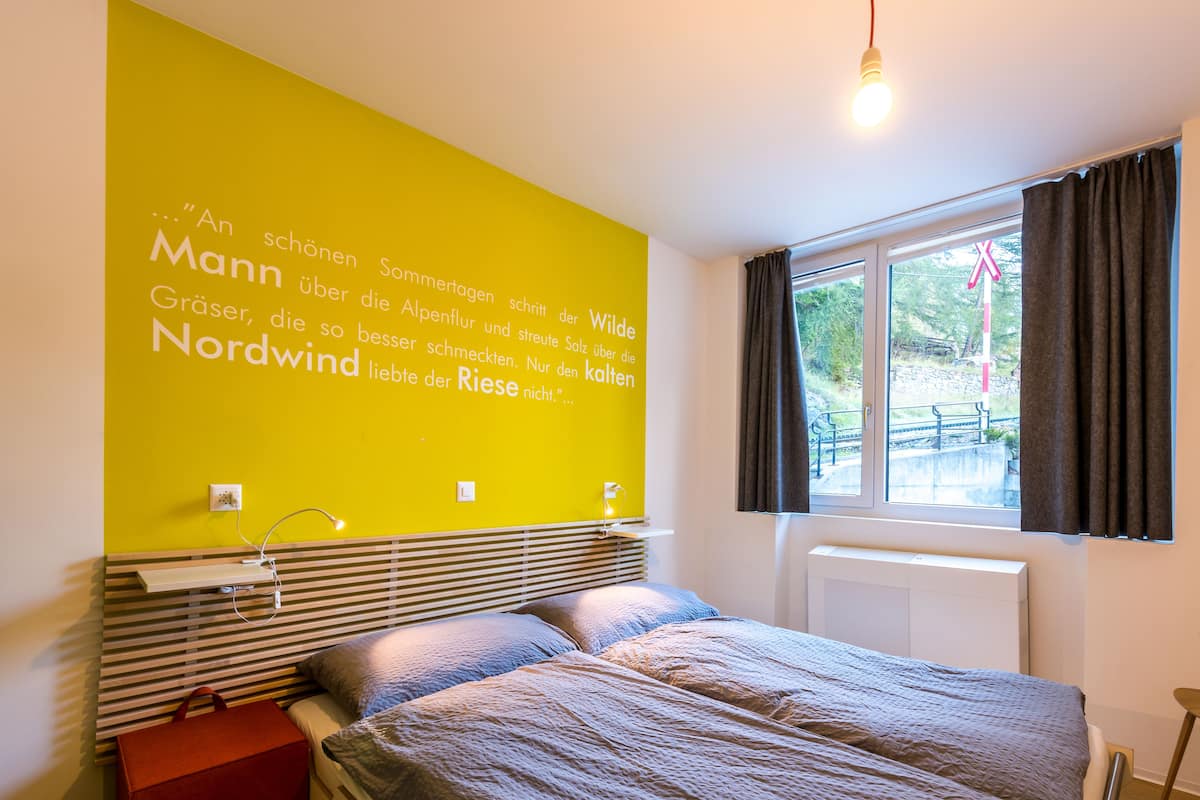 A bedroom with a bed, a bedside table, a lamp, a window with curtains, and a wall with text.