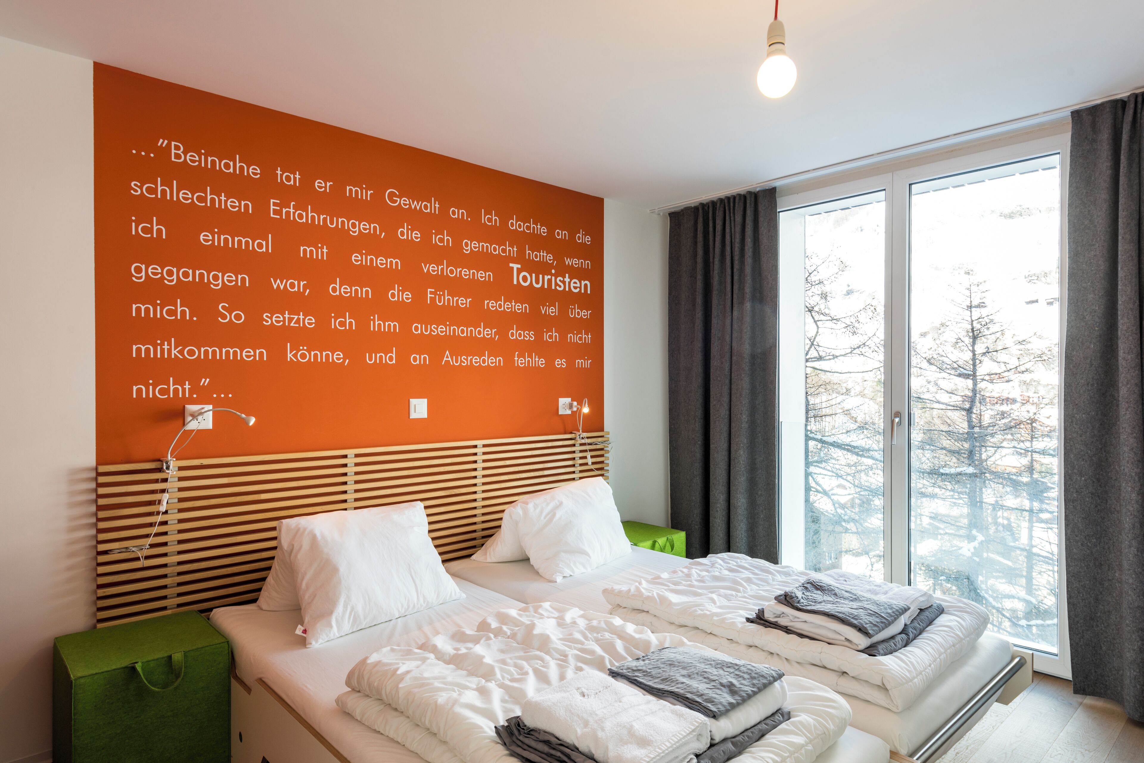 A hotel room with two beds, a large window with curtains, and a wall with a quote in German.