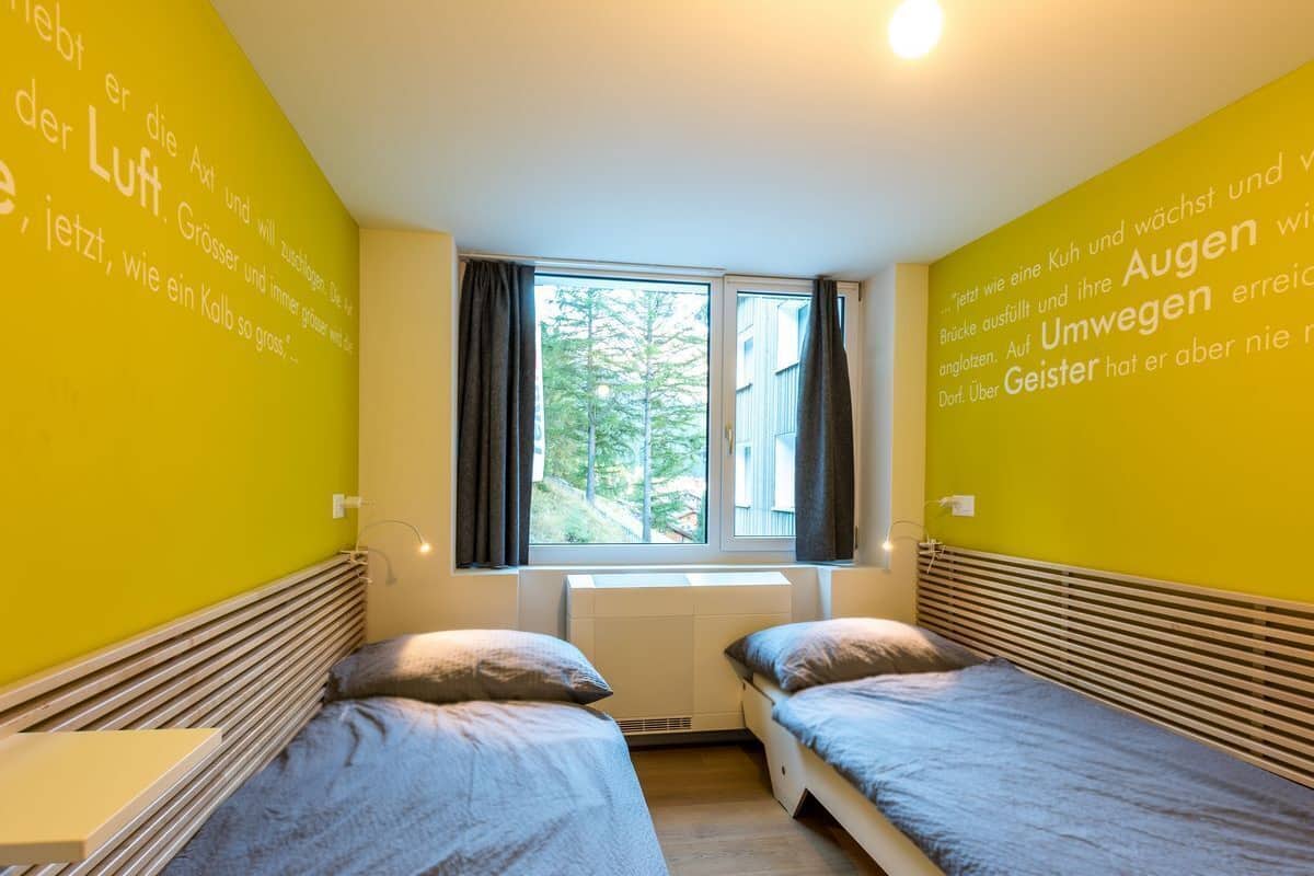 A room with two beds, a window with curtains, and a wall with text in a foreign language.