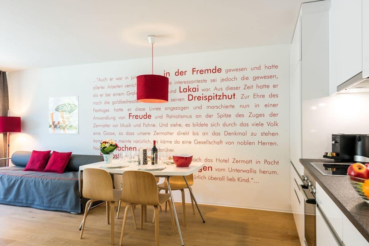 A modern kitchen with a dining area, featuring a red pendant light, a white wall with text, and a cozy seating arrangement.
