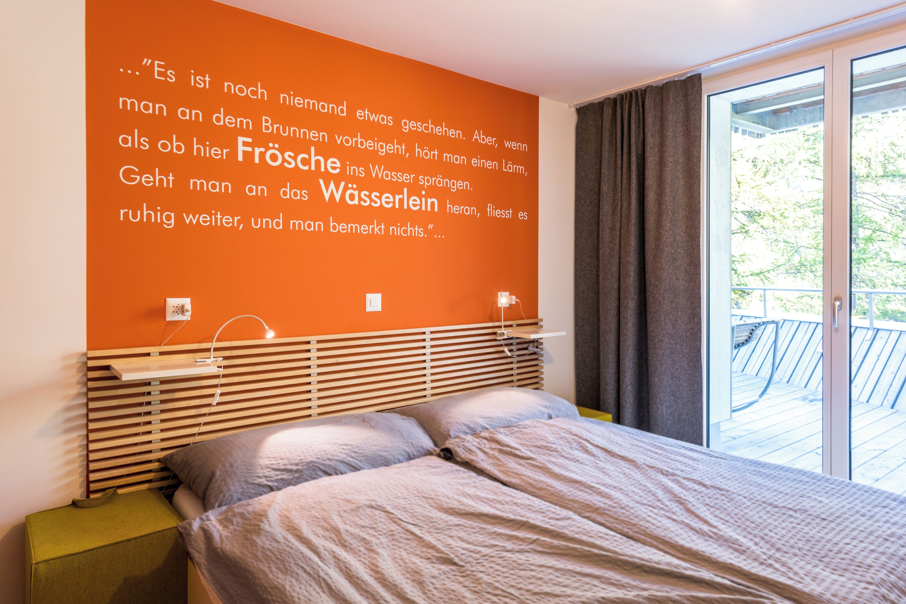 Ein Schlafzimmer mit großem Fenster, einem Bett, einem Nachttisch und einer Wand mit einem deutschen Zitat.