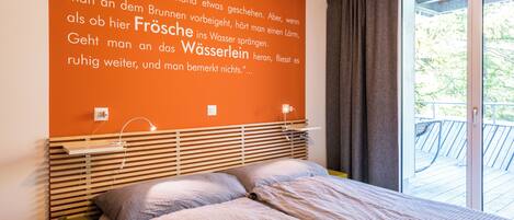 Ein Schlafzimmer mit großem Fenster, einem Bett, einem Nachttisch und einer Wand mit einem deutschen Zitat.