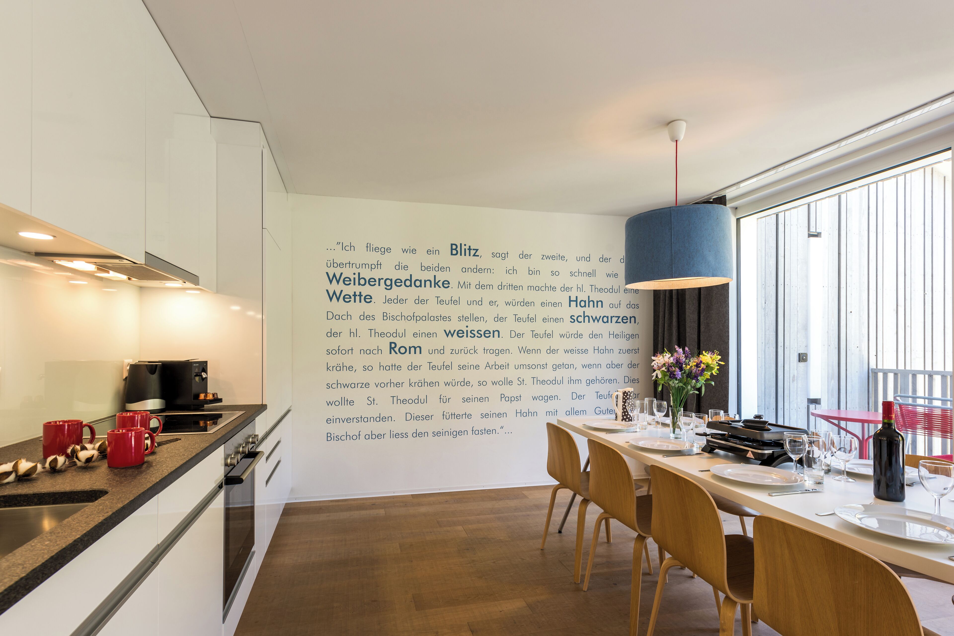 A modern kitchen with a dining area, featuring a long table set for dining, a coffee machine, and a wall with text.