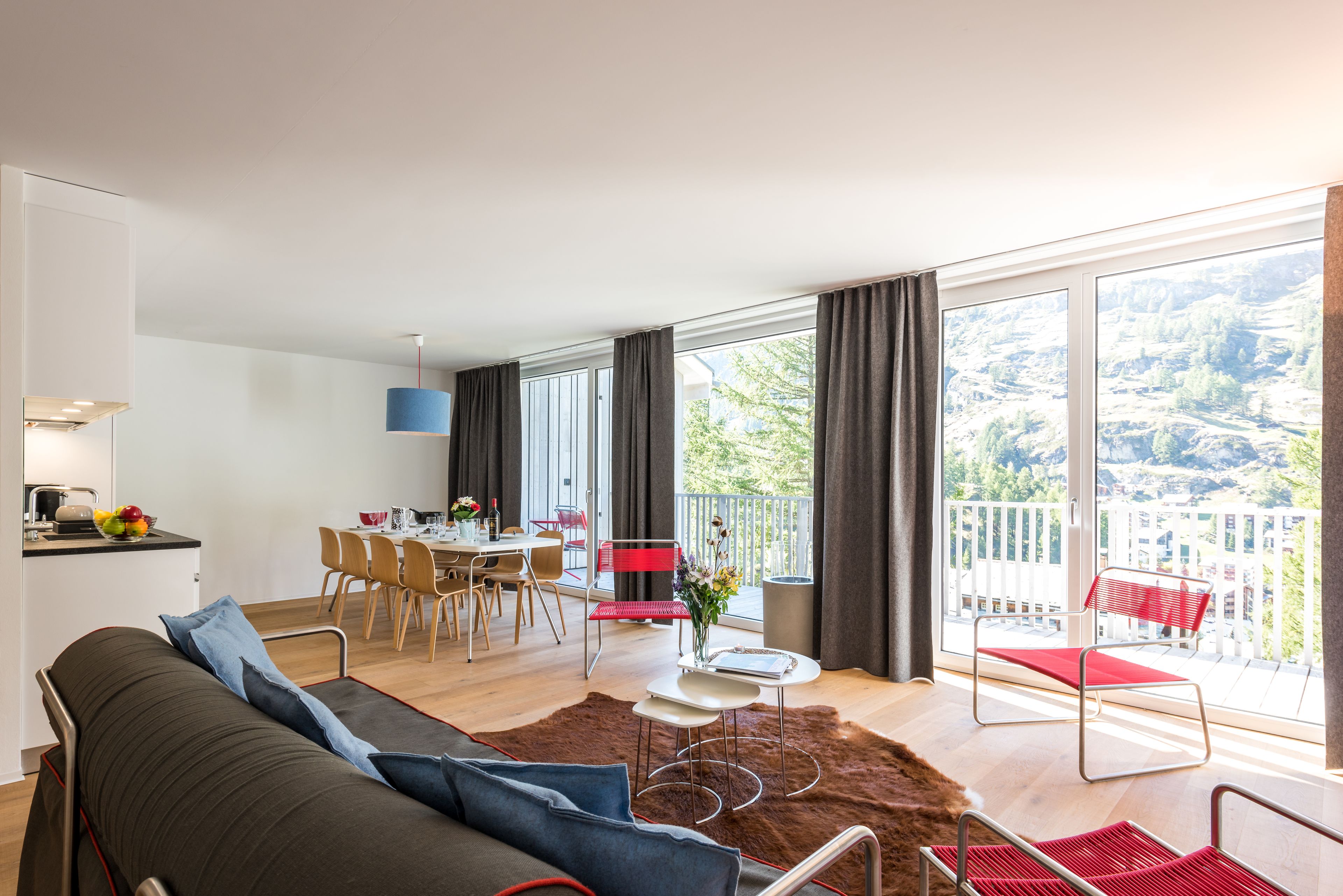 A modern living room with a sofa, a dining area with chairs, a small table with a vase, and a balcony with a view of the outdoors.
