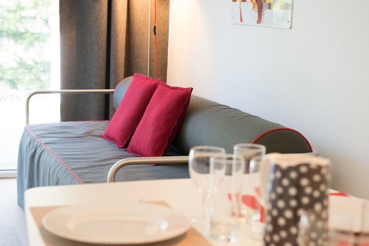 A neatly arranged hotel room with a bed, a table set with plates and glasses, and a view of the outdoors through the window.