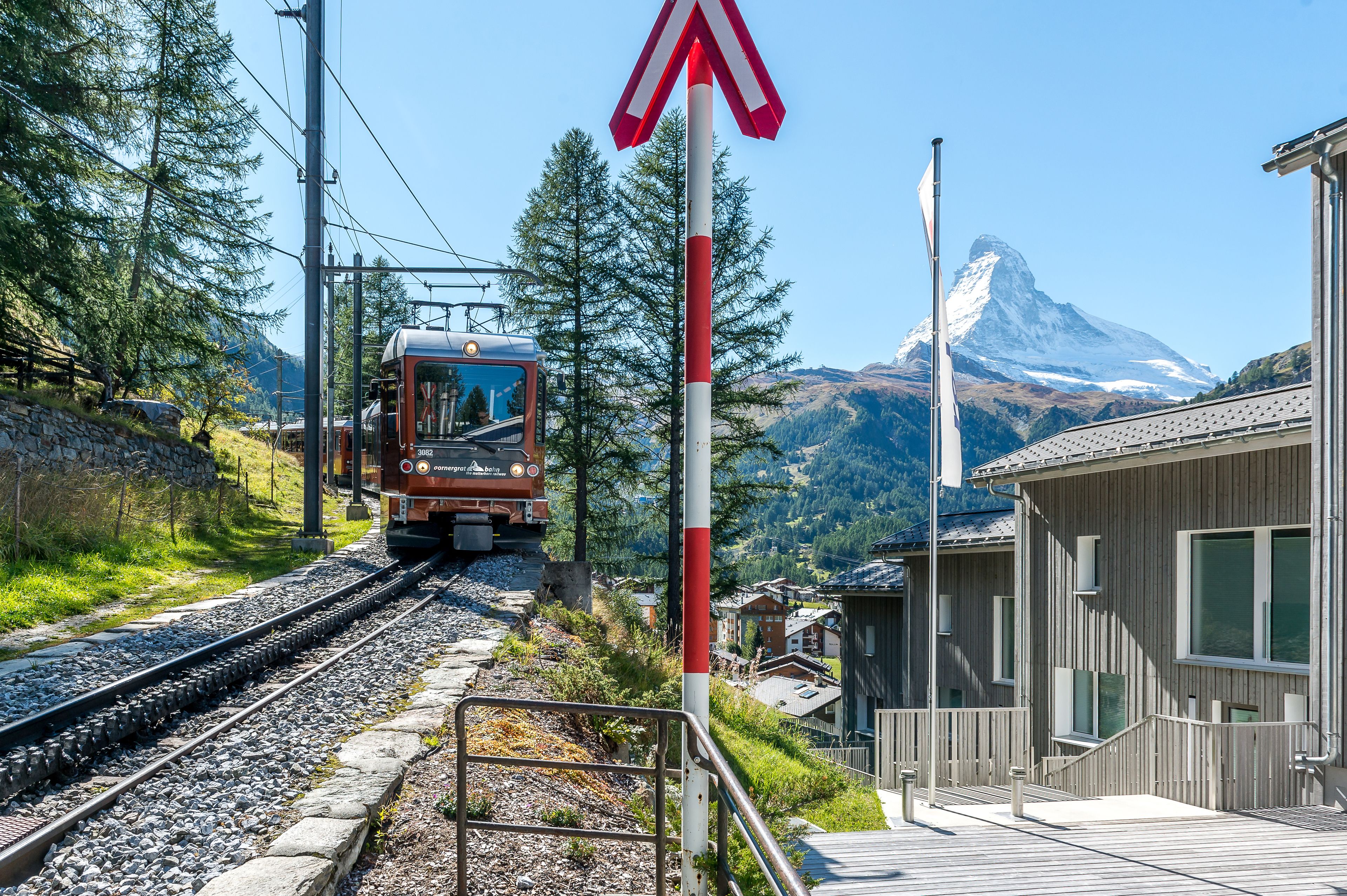 A train on tracks with a red and white crossing sign, a wooden building, and a mountain in the background.