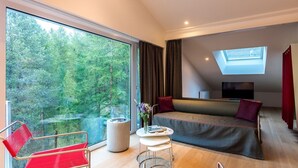 Une chambre d’hôtel moderne avec une grande fenêtre, un canapé, un fauteuil rouge, une petite table sur laquelle est posé un vase de fleurs et un téléviseur.