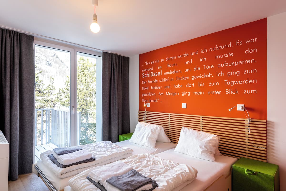 A modern hotel room with a large bed, a view of trees through the window, and a wall with text in a foreign language.