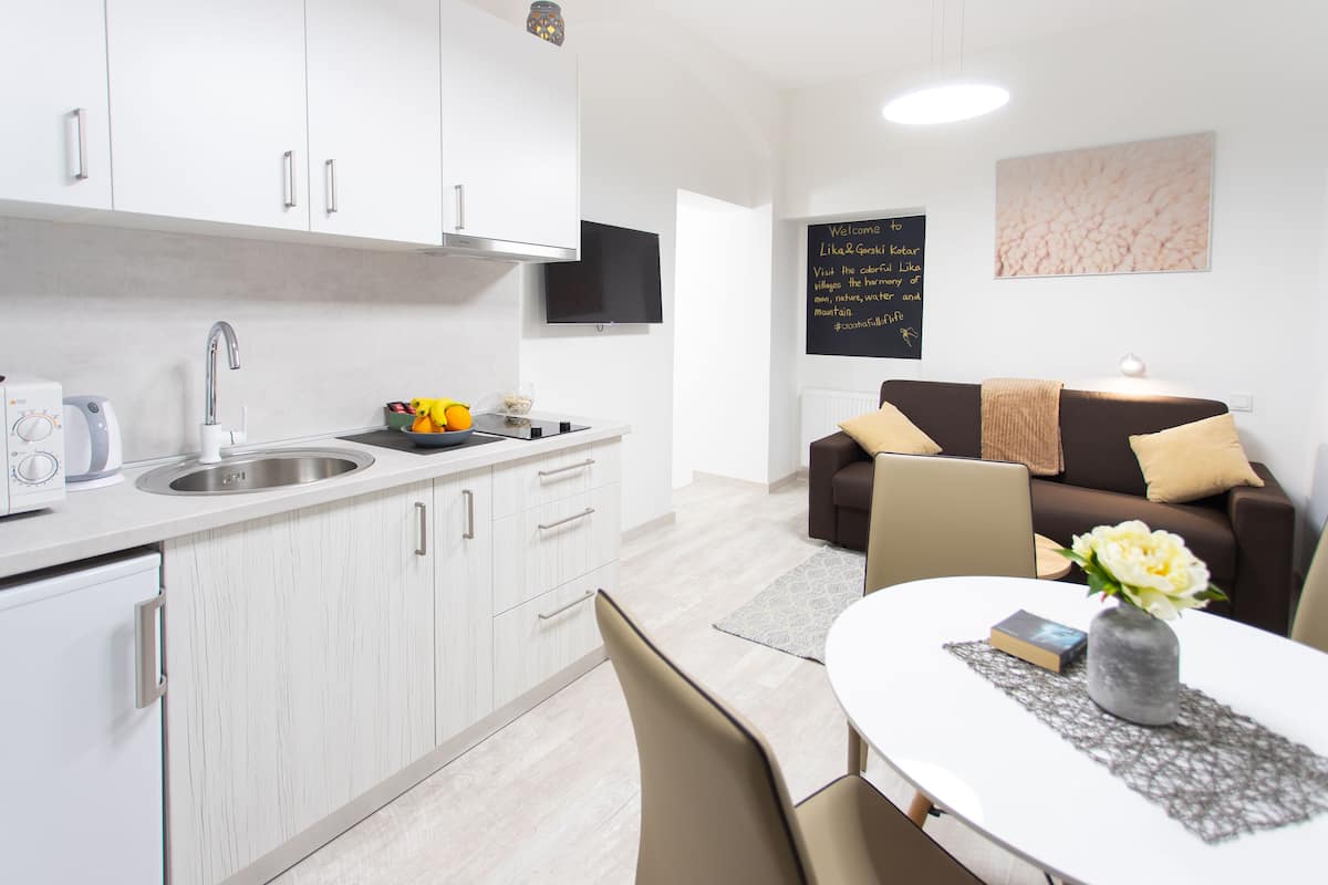 A modern kitchen with white cabinets, a built-in oven, and a sink. There is a dining area with a table, chairs, and a vase with flowers. A brown sofa with cushions is in the background.