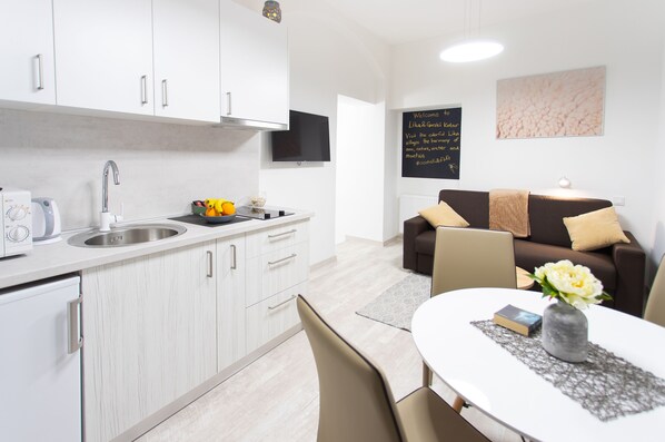 A modern kitchen with white cabinets, a built-in oven, and a sink. There is a dining area with a table, chairs, and a vase with flowers. A brown sofa with cushions is in the background.