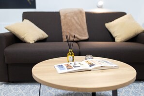 A modern living room with a dark brown sofa, two beige cushions, a round wooden coffee table with magazines and a bottle of wine.