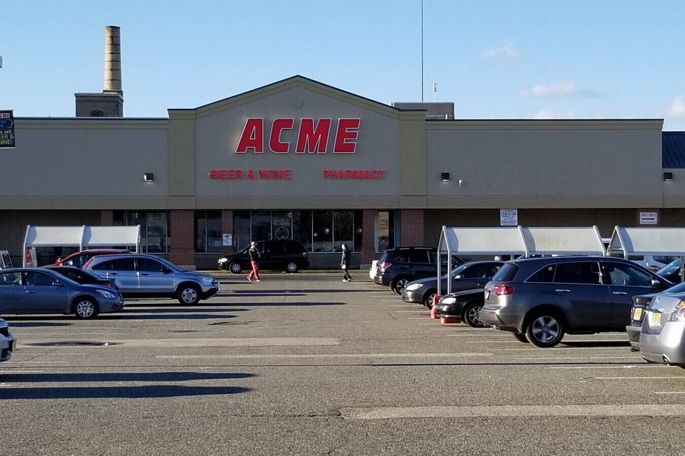 Une pharmacie avec une enseigne indiquant « PHARMACIE ACME – BIÈRES ET VINS » et un parking où sont garées plusieurs voitures.