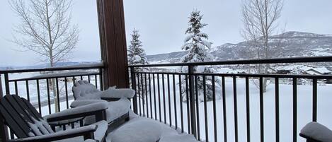 A snow-covered deck with outdoor seating and a view of snow-covered trees and mountains.
