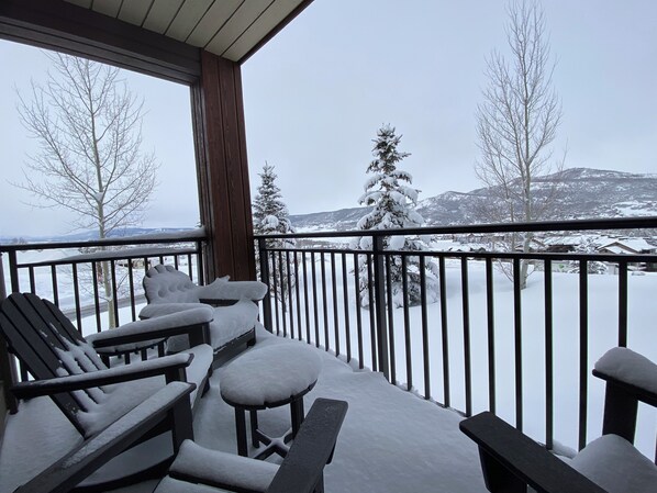 A snow-covered deck with outdoor seating and a view of snow-covered trees and mountains.