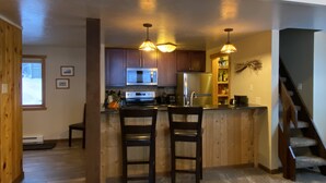 A kitchen with wooden cabinets, a microwave, and a refrigerator. There are two bar stools and pendant lights above the counter.