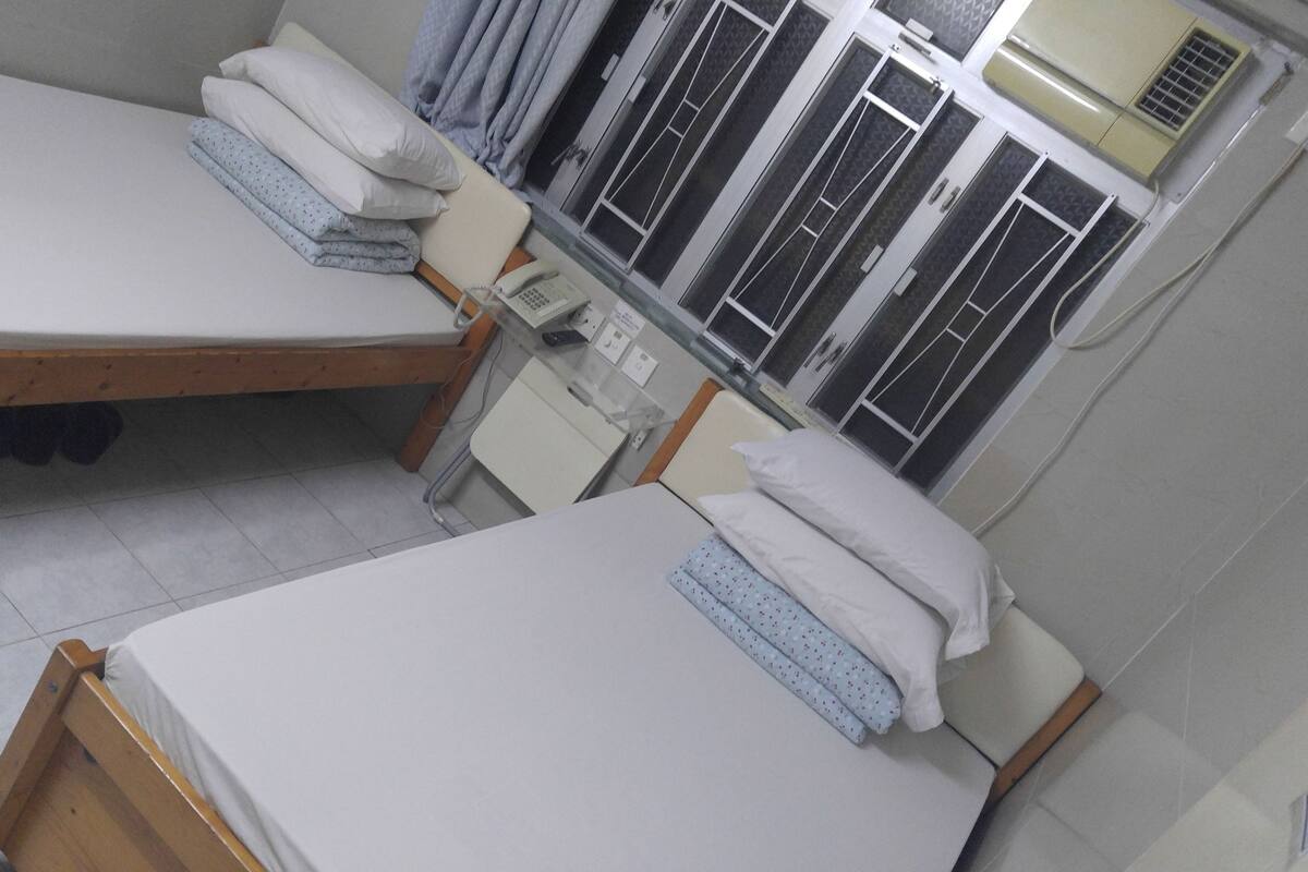 Een kamer met twee enkele bedden, een klein bureau met telefoon en een raam met metalen tralies.