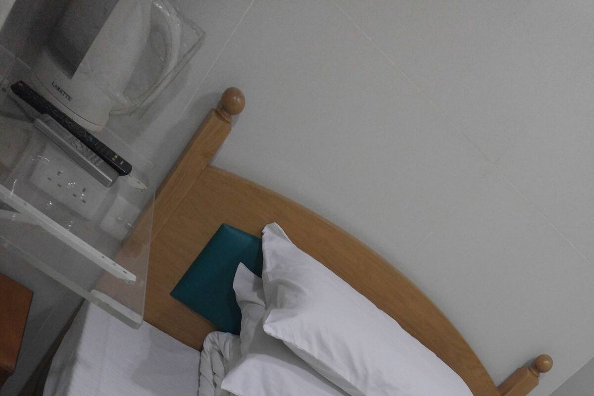Een keurig opgemaakt bed met wit beddengoed en kussens, een houten hoofdbord en een klein nachtkastje met een lamp en afstandsbediening.