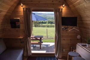 Una cabaña acogedora con interior de madera, una cama, un escritorio, un televisor y vistas a un paisaje cubierto de césped a través de un gran ventanal.