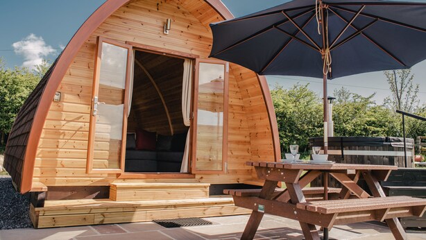 Een houten A-frame constructie met een open deur, een picknicktafel en een parasol.