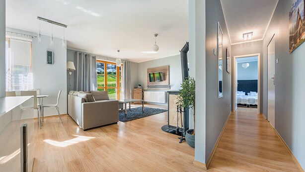 Een moderne woonkamer met een bank, een eettafel en stoelen, een keuken met een fornuis, en een hal die leidt naar een slaapkamer met een bed.