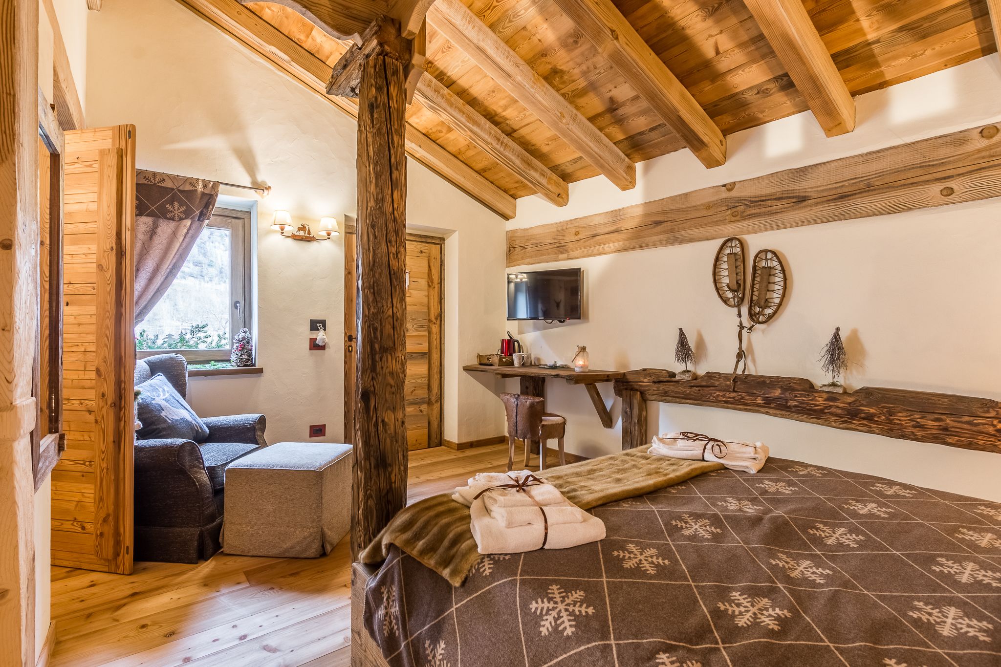 Una habitación acogedora con techo de madera, una cama con colcha estampada, una silla, una mesita con televisor y una ventana con cortinas.