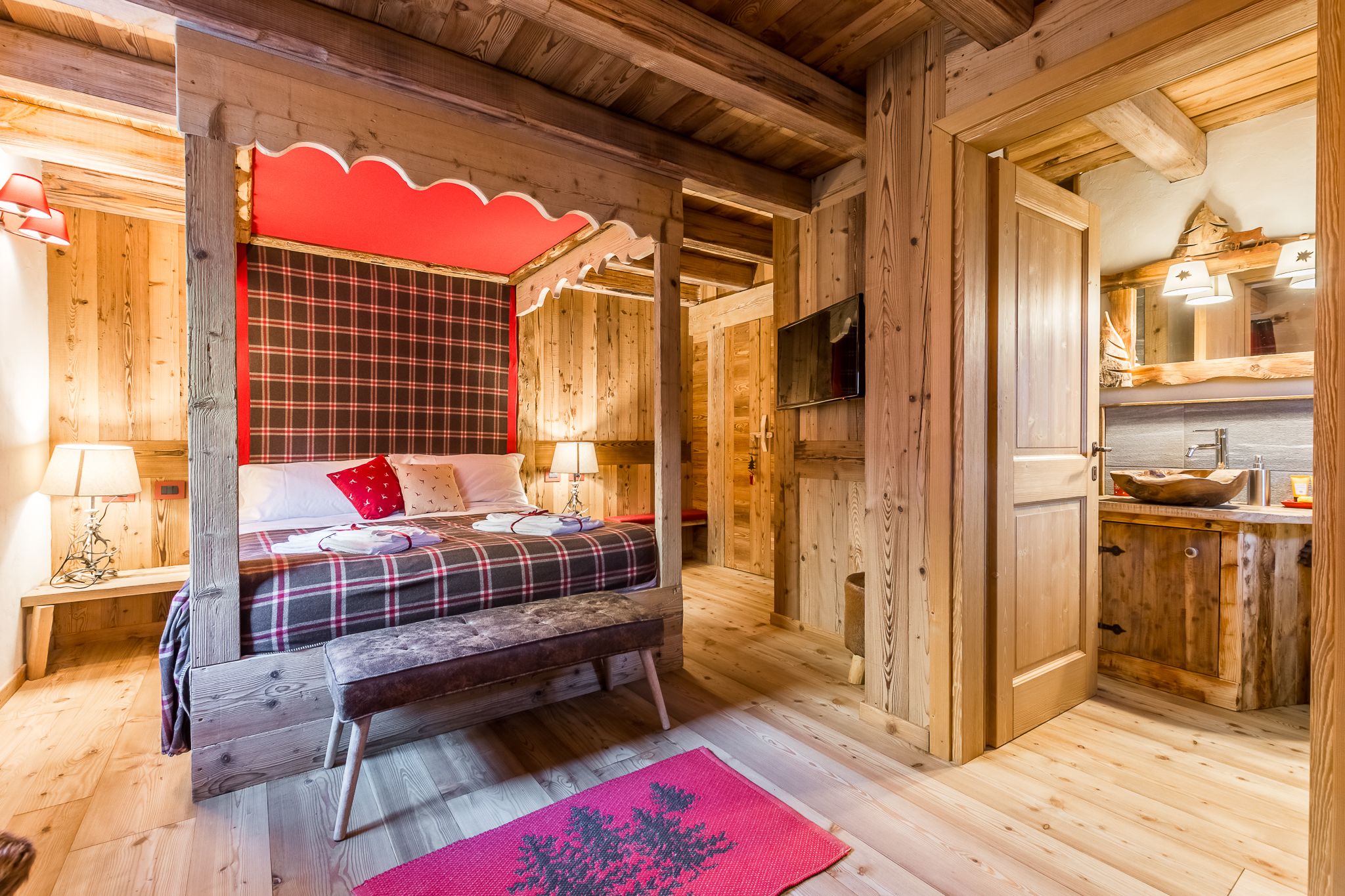 Una cabaña de madera con una cama, un banco, un cabecero rojo a cuadros, un televisor y un gabinete de madera con un lavamanos.