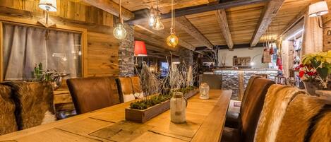 Un rústico comedor de madera con una larga mesa, sillas de cuero, lámparas colgantes y un fondo de pared de piedra.